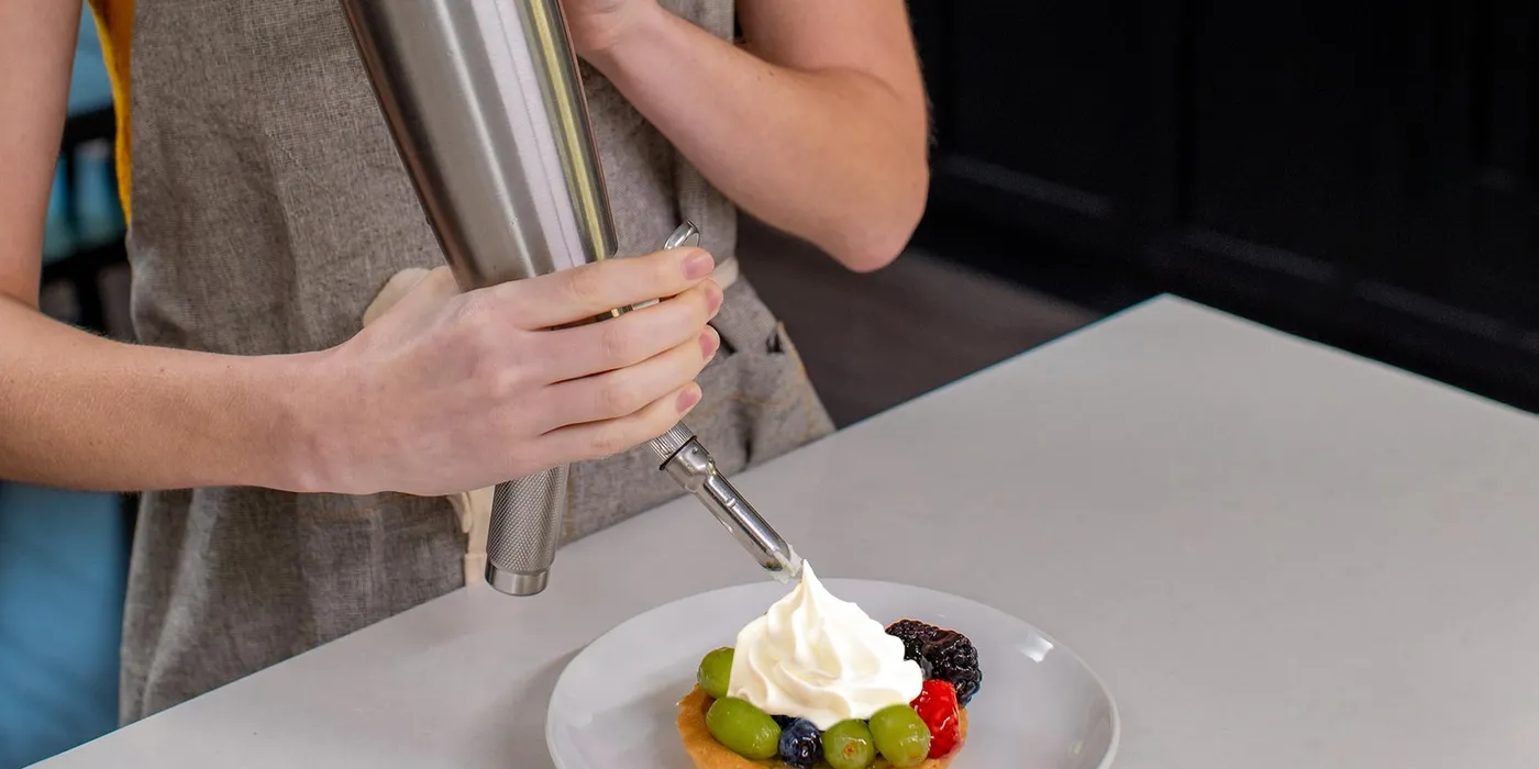 Piping fresh whipped cream onto a fruit tart with a stainless steel whipped cream dispenser